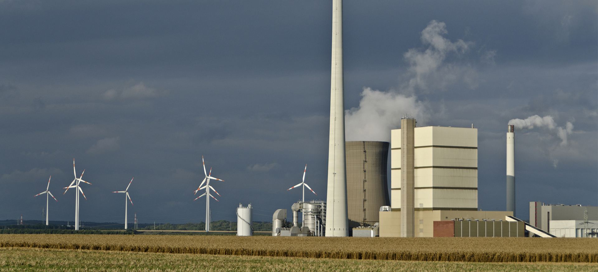 Kraftwerk mit rauchenden Schornsteinen und Windkraftanlagen vor dunkelgrauem Himmel auf einem Feld