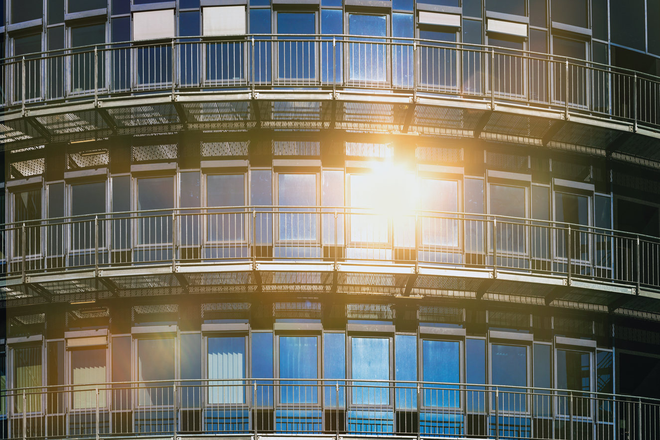 The picture shows the glass frontage of a house reflecting the sun. Some of the windows are part-shaded by means of external roller shutters.