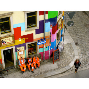 Drei Männer in den orangenen Anzügen der Müllabfuhr sitzen auf einer Bank vor einem bunt gestrichenen Eckhaus und trinken Kaffee