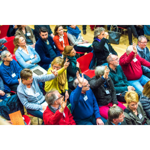 Es sind viele Menschen in 5 Stuhlreihen zu sehen. Sie sind von oben fotografiert. Es ist ein fröhliches Bild, die Menschen wirken sehr aufgeweckt und tragen bunte Kleidung. Die Menschen melden sich und beteiligen sich rege.