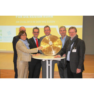 Hinter einem Stehtisch stehen sechs Personen. Sie posieren für ein Foto. Eine Frau hält eine große goldene Klangplatte und einen hölzernen Hammer,  mit dem sie auf die Platte schlägt, um einen Gong zu erzeugen. 