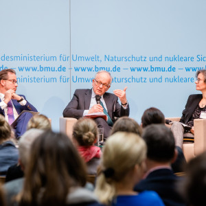 Podiumsdiskussion vor hellblauem Hintergrund, auf dem der Veranstaltungsort geschrieben steht.