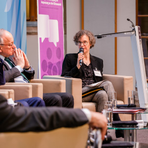 Podiumsdiskussion aus einer anderen Perspektive. Im Hintergrund steht ein Rollup in lila Farbtönen mit Veranstaltungstitel. Die Moderatorin spricht in das Mikrofon.