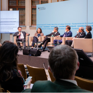 Podiumsdiskussion aus einer anderen Perspektive. Im Hintergrund steht eine Tafel, auf der die Podiumsbeteiligten verzeichnet sind.