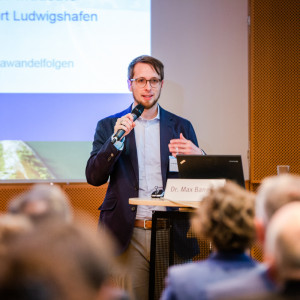 Vortragender steht mit Mikrofon in der Hand am Stehpult. Im Hintergrund ist seine Präsentation abgebildet.