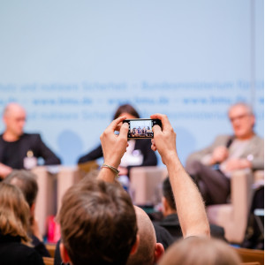 Ein Teilnehmer hält ein Smartphone in die Luft in Richtung Podium und macht ein Foto.