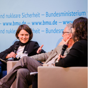 Podiumsdiskussion vor dem hellblauem Hintergrund, auf dem der Veranstaltungsort geschrieben steht. Eine Teilnehmende hält ein Handmikrofon und spricht angeregt zu den anderen Podiumsteilnehmenden.