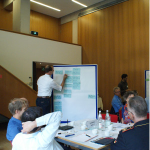 Ein Teilnehmender schreibt etwas auf eine Präsentationtswand, die mit blauen Moderationskarten bestückt ist. Im Raum sitzen alle Teilnehmenden an ihren Gruppenarbeitstischen.