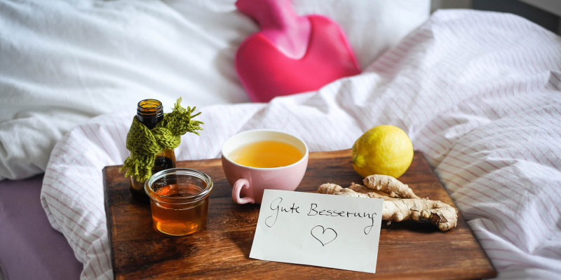 Foto Bett mit Wärmflasche und Tablett, darauf Honig, Tee, Ingwer, Zitrone und Schild mit „Gute Besserung“