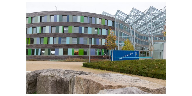 Hauptgebäudes des UBA in Dessau-Roßlau, davor steht ein Container mit dem Schriftzug "Waterlines.eu". Ein blauer Laser zeigt den Wasserpegel an.