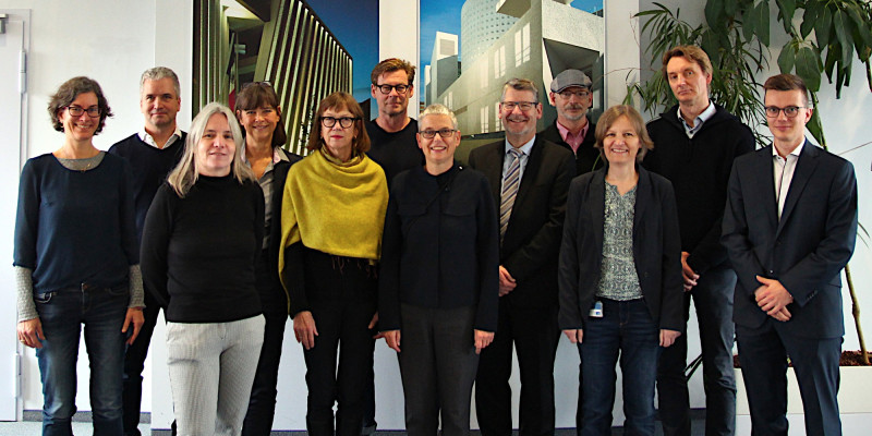 Gruppenfoto mit zwölf Personen in zwei Reihen vor einer Wand mit zwei Architekturbildern.