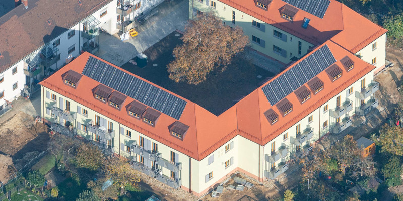 Luftbild eines Wohngebäudes mit drei Flügeln in einem U. Zwei Vollgeschosse und ein Walmdach mit mehreren Gauben und PV-Anlagen. Im Innenhof ein großer Baum, in der Umgebung Kleingärten. Vor der Öffnung des U ein linienförmiger Wohnbau ähnlicher Kubatur.