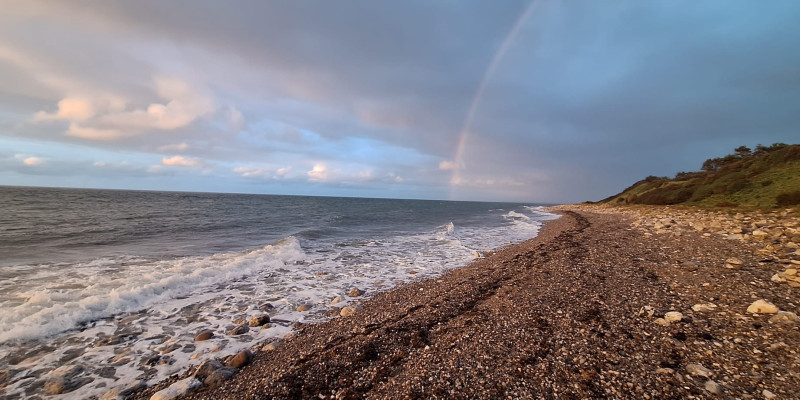 Wellen der Ostsee schlagen an einen Steinstrand, der in grüne Hügel übergeht. Der Himmel ist über den Hügeln graublau aber über dem Meer hellblau mit weißen Wolken. Ein Regenbogen beginnt über dem Meer.  