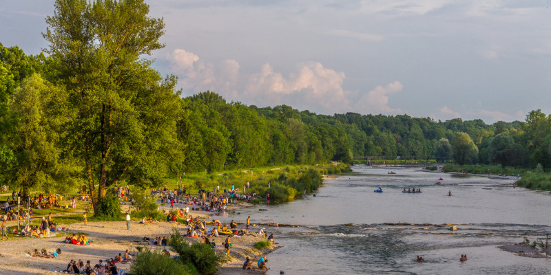 Foto: Ein renaturierter Abschnitt der Isar bei München mit zahlreichen Badegästen am Kiesstrand und im Wasser.