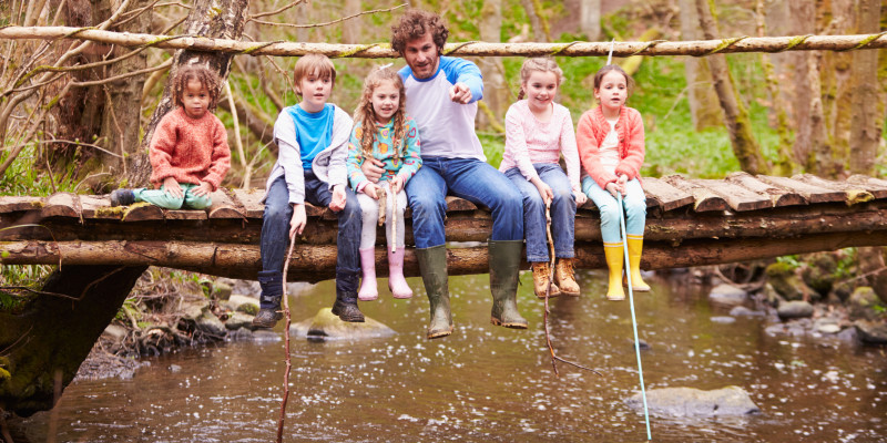 Foto: Mann sitzt mit fünf Kindern auf einer Holzbrücke, die über einen Waldbach führt. Der Mann zeigt auf etwas im Bach. Ein Kind hat einen Kescher in der Hand.