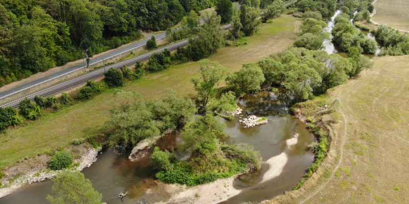 Luftbild der Fulda mit großzügiger Aufweitung und Kiesbänken, die teilweise mit Vegetation bedeckt sind. Links ist eine Straße zu erkennen, rechts grenzt Grünland an das Gewässer an.