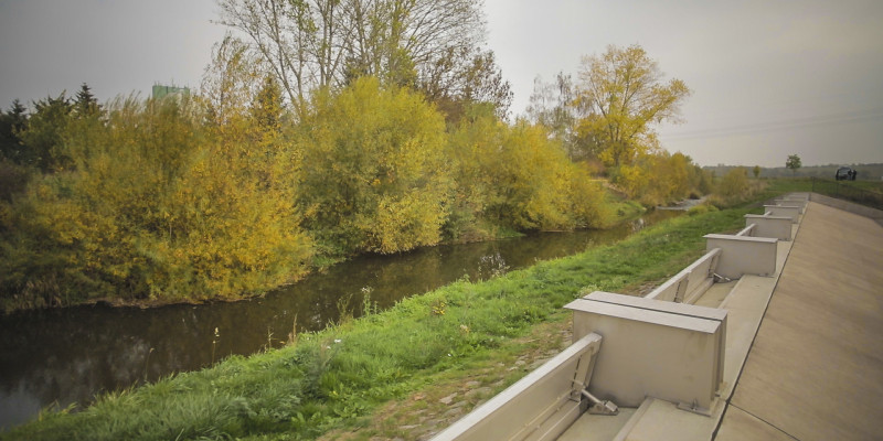 Foto: Ein Klappenwehr aus Beton und Stahl entlang der Helme. Das Ufer im Bereich vor dem Klappenwehr ist mit Gräsern bedeckt. Die andere Uferseite ist naturnah mit Gehölzen bestanden.