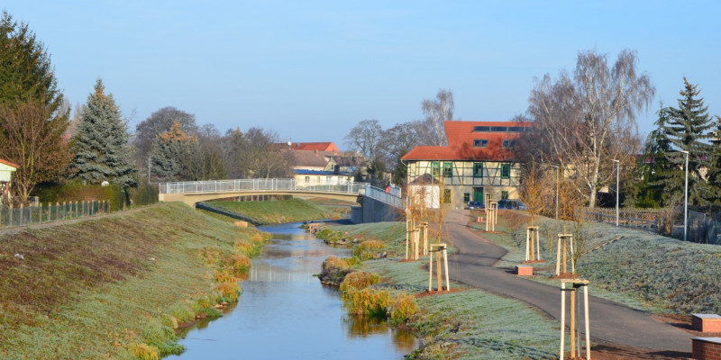 Foto: Die renaturierte Helme in Sundhausen. Die Breite der Helme variiert leicht mit weiten und engeren Abschnitten. Entlang der Ufer sind kleinere Gewächse zu erkennen. In der Umgebung finden sich Häuser und befestigte Wege.