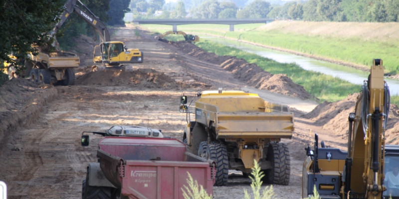 Foto: Zahlreiche Kipplaster helfen bei der Uferabflachung durch zwei Bagger. Im Hintergrund liegt das zu renaturierende Gewässer.