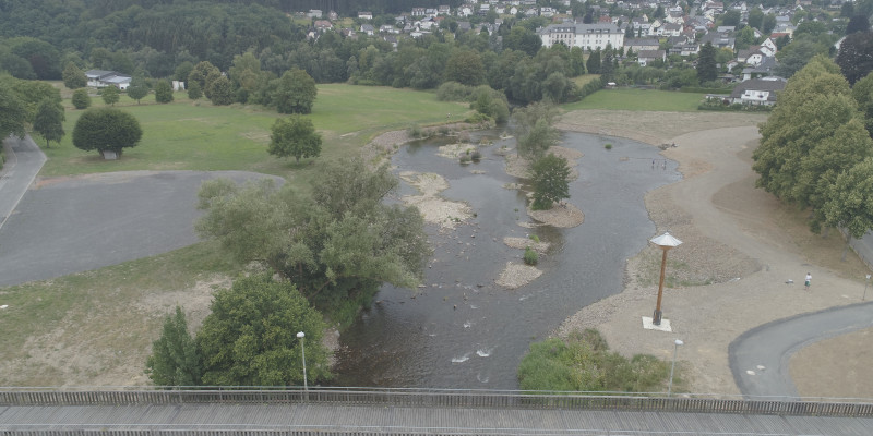 Luftaufnahme eines renaturierten Gewässers. Unter einer Brücke strömt ein breiter Fluss mit vielen kleinen Inseln aus Kies und Vegetation. An das Gewässer grenzt ein kleiner, unbefestigter Weg an und im Hintergrund ist eine größere Siedlung erkennbar.