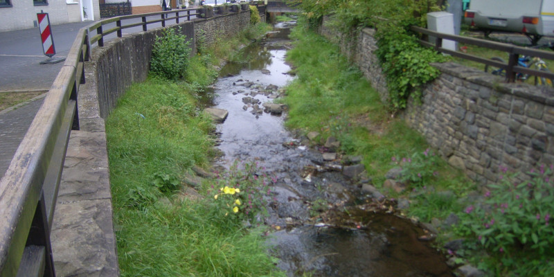 Foto: Ein Bach mit hohen Ufermauern und angrenzenden Häusern und Straßen in Ortslage. Die Gewässersohle ist offen, mit Kies bedeckt und stellenweise bewachsen. Der Bach verläuft leicht schwingend im Kastenprofil.