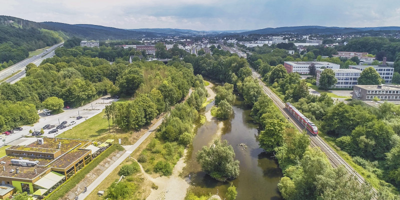 Luftbildaufnahme der renaturierten Ruhr, die umgeben ist von Häusern, Straßen und einer Bahnlinie. Ein Ausflugslokal bietet direkten Blick auf die Renaturierung. Im Fluss selbst liegen Kiesbänke und Totholz. Der Uferbereich ist mit zahlreichen Gehölzen bewachsen.