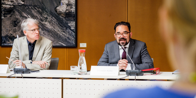 Das Bild zeigt Christian Kühn, den Parlamentarischen Staatssekretär des Bundesministeriums für Umwelt, Naturschutz, nukleare Sicherheit und Verbraucherschutz . Dieser sitzt an einem Tisch und begrüßt die Jury