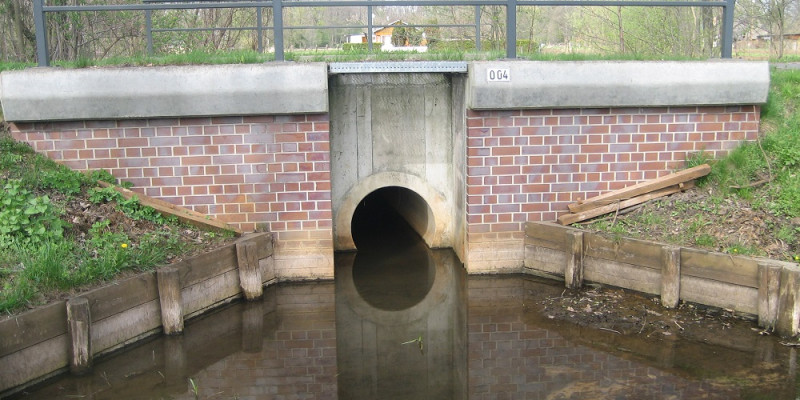 Auf dem Foto ist die Verrohrung eines Bachs unter einer Brücke dargestellt