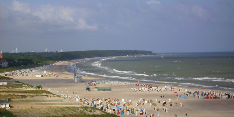 Blick auf Strand mit Strandkörben und touristischer Infrastruktur, im Wasser Kite- und Windsurfer. 