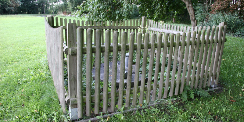 Umzäunter Brunnen mit Holzlatten auf grüner Wiese in Großaufnahme.