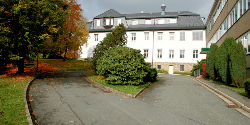 vierstöckiger weißer Altbau mit einer Auffahrt durch eine Grünanlage, rechts ein dreistöckiger älterer Neubau