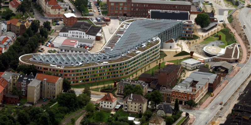  The UBA building in Dessau-Roßlau from above. The building is elongated, with a façade of wood and coloured glass slabs and slanted solar cells covering the entire surface of the flat roof