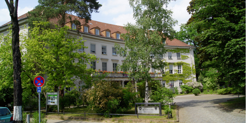 Three-storey historic building with entrance portal, with balcony and flower boxes above, gabled roof with many small dormers. At the front a square with trees, bushes, a drive and sign which reads "Umweltbundesamt"