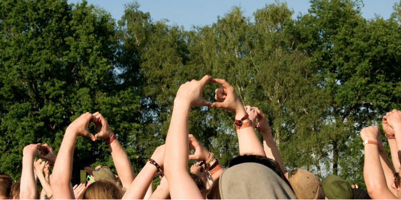 Menschen auf einem Musikfestival im Grünen strecken die Arme in die Höhe und formen mit ihren Händen ein Herz