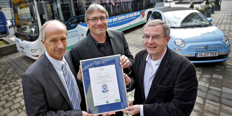 drei Männer halten eine Urkunde mit dem Umweltzeichen "Blauer Engel", im Hintergrund steht ein Linienbus