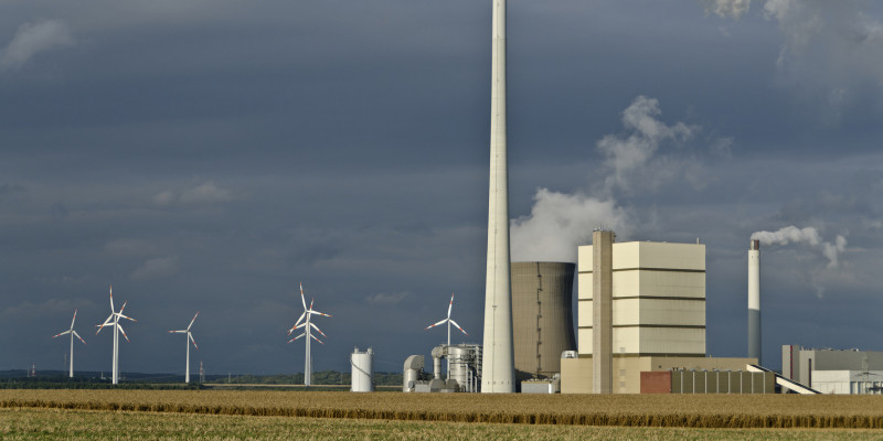 Kraftwerk mit rauchenden Schornsteinen und Windkraftanlagen vor dunkelgrauem Himmel auf einem Feld