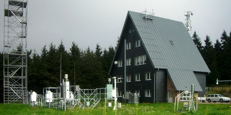 Small, four-storey building with dark brown wooden façade and gabled roof, with measuring devices on a field in front