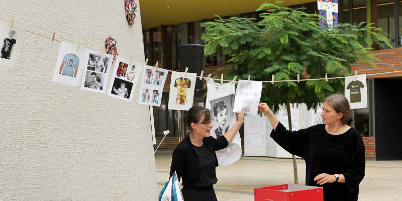 Kunstausstellung: Zwei Frauen stehen vor einer Wäscheleine und einem Bügelbrett und hängen Informationen für die Besucher an die Leine