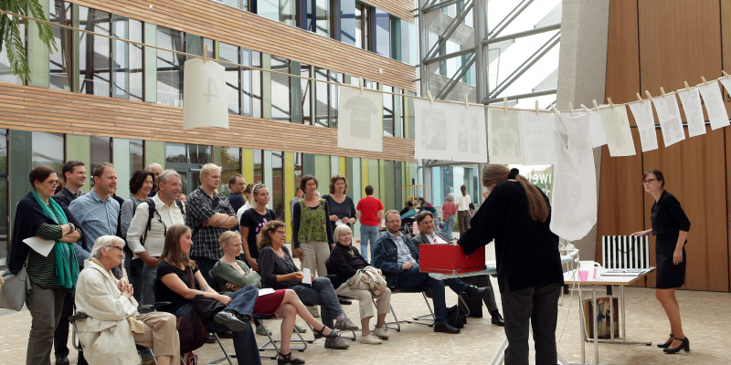 Kunstausstellung: Zwei Frauen stehen vor einer Wäscheleine und einem Bügelbrett und hängen Informationen für die Besucher an die Leine, davor Publikum