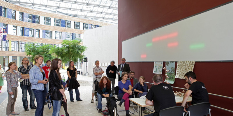 Vier Personen sitzen an einem Tisch, darum Ausstellungsbesucher