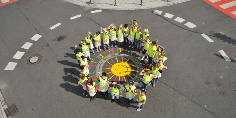 Kinder stehen jubelnd um einen Gullideckel, den sie bunt mit einer Sonne bemalt haben