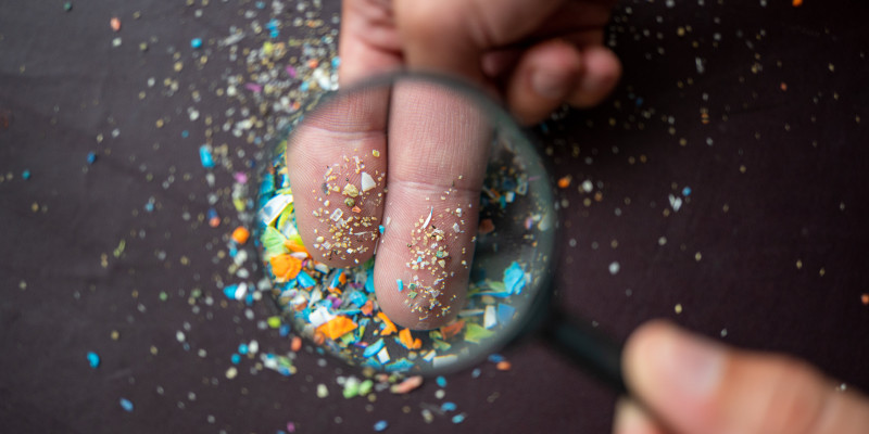 Blick durch eine Lupe auf kleine bunte Mikroplastikteile, die auf zwei Fingern gehalten werden.
