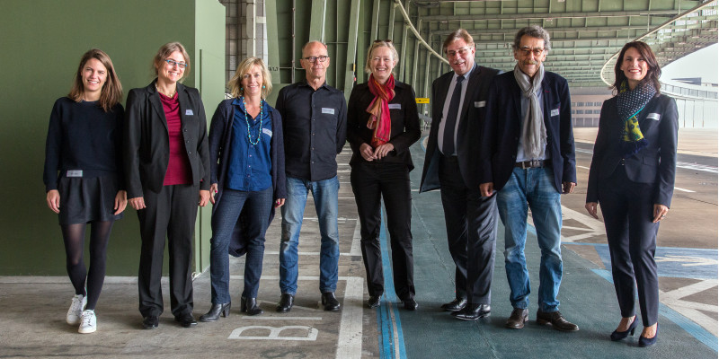 Zu sehen sind die acht Personen der Jury auf dem Rollfeld des ehemaligen Berliner Flughafens in Tempelhof.