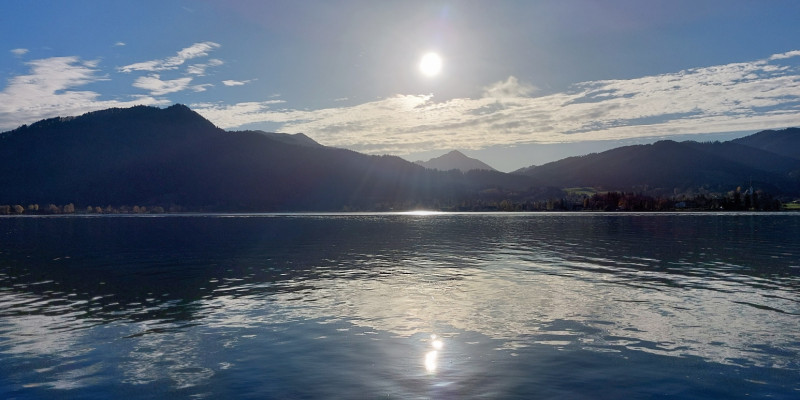 Das Bild zeigt einen Gebirgssee bei gleißendem Sonnenlicht. Im Hintergrund ist eine Bergkette zu sehen.