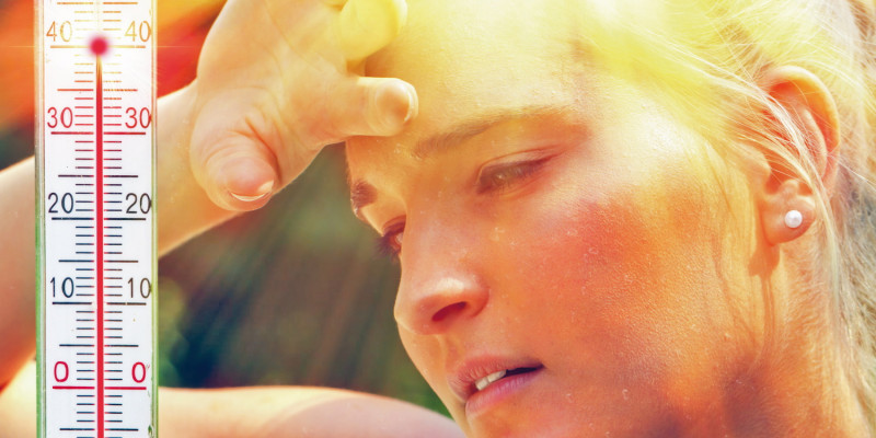 Das Bild zeigt das Gesicht einer Frau mit Schweißperlen in der Sommerhitze. Die Frau hält die Hand an die Stirn. Links im Bild ist ein Thermometer abgebildet, das 40 °C Lufttemperatur anzeigt.