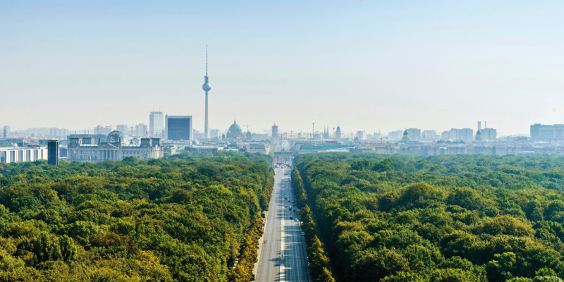 Das Bild zeigt die Straße des 17. Juni in Berlin aus der Luftperspektive. Rechts und links der Straße sieht man das Blätterdach der Bäume des Großen Tiergartens. Am Bildhorizont ist die Silouhette der Stadt zu erkennen, unter anderem mit Reichstagsgebäude, Fernsehturm, Dom und Brandenburger Tor.