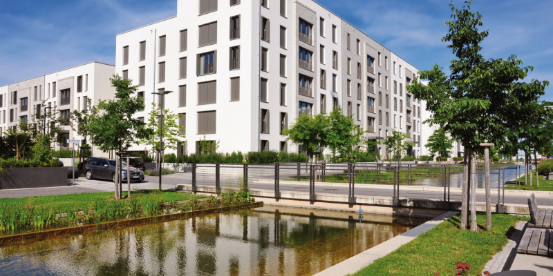 Das Bild zeigt einen künstlichen Wasserlauf, der in einem neu entwickelten Quartier an einem Geschosswohnungsbau vorbeifließt. Eine Straße quert den Wasserlauf. Der Wasserlauf ist von einem schmalen Grünstreifen mit Rasen, jungen Bäumen und Blumenbeeten eingefasst. Bänke bieten Sitzmöglichkeiten.