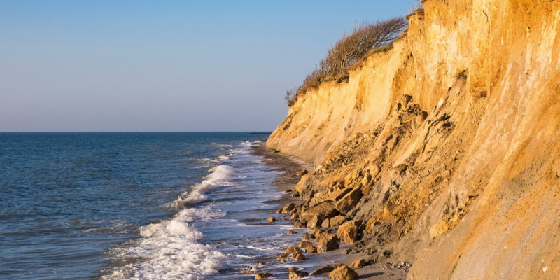 Das Bild zeigt die hellbraune Steilküste bei Ahrenshoop an der Ostsee. Am Fuß des Kliffs ist das Meer und die Gischt einer Welle zu sehen. An einem Küstenabschnitt befindet sich lockeres Material und Gestein, das sich von der Kliffoberkante an den Klifffuß verlagert hat.