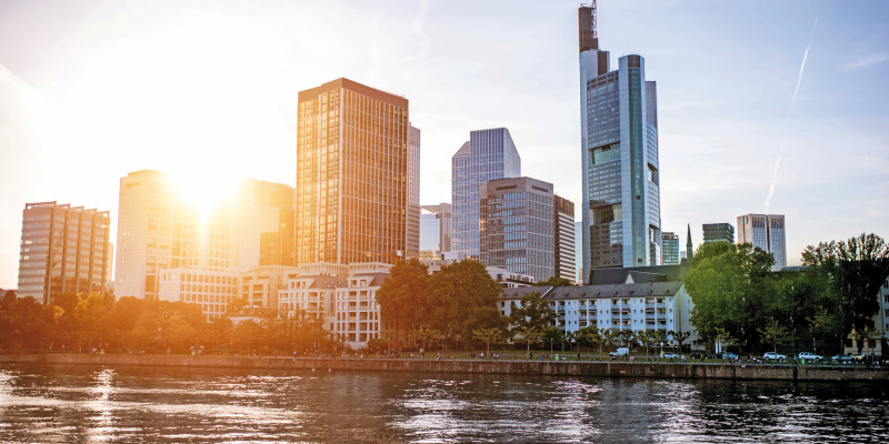 Das Bild zeigt einen Ausschnitt der Skyline von Frankfurt am Main, beschienen von einer tief stehenden Sonne. Im Vordergrund ist der Fluss zu sehen.