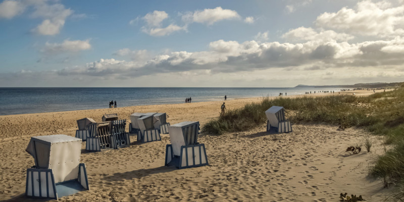 Das Bild zeigt einen Sandstrand an der Ostsee mit Dünen und Strandkörben. Die der oder dem Betrachtenden zugewandten Strandkörbe sind versperrt. Am Horizont ist das Meer zu sehen. An der Wasserlinie sind einzelne Personen und Personengruppen zu erkennen, sonst ist der Stand leer. Der Himmel ist in Teilen bewölkt.